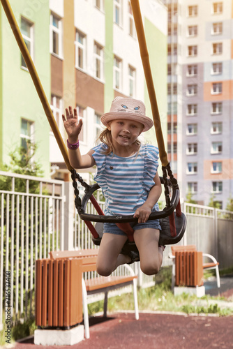 Happy little toddler 4 year girl in summer hat having fun on swing in playground in sunny day, looking at camera and say hello