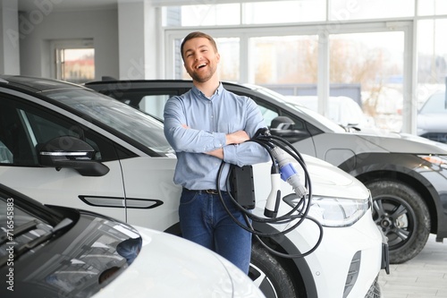 Handsome business man holding charging cable for electric car. Caucasian male stands near electric auto in dealership © Serhii