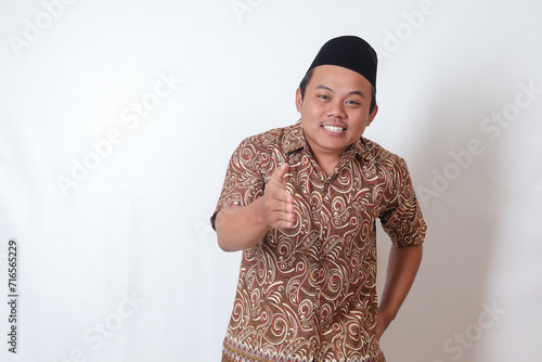 Portrait of excited Asian man wearing batik shirt and songkok reaching out his hand for a hand shake, welcoming someone. Isolated image on gray background photo