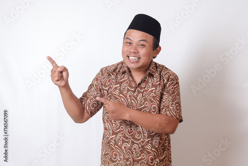 Portrait of excited Asian man wearing batik shirt and songkok smiling and looking at the camera pointing with two hands and fingers to the side. Isolated image on gray background