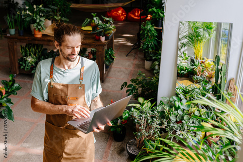 Caucasian guy wearing apron working with laptop in a home indoor. Man working with laptop or computer in cozy room with greenery plants.