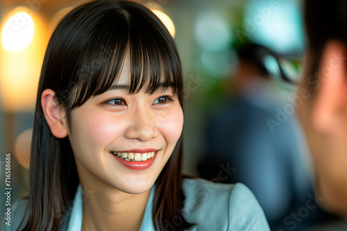 corporate photo, close-up and clear focus on a young Japanese female office worker with minimal makeup, hair with subtle bangs, warmly smiling and talking to a blurred man out of frame