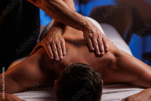 Physiotherapist massaging male patient with damaged shoulder muscle, treating sports injuries. Relaxing professional shoulder massage in cozy atmosphere, reboot on the weekend, end of difficult week.