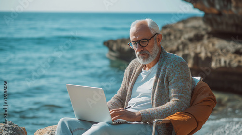 mature man with laptop working outdoors while sitting on bench at seaside.