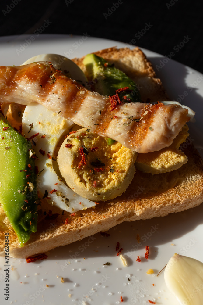 A healthy breakfast of toast topped with boiled egg, avocado, and grilled chicken breast, served on a white plate bathed in sunlight from a window.