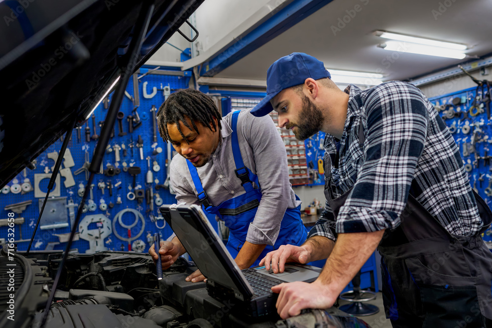 Dynamic duo at work: Mechanic colleagues dive into car diagnostics with a computer, complemented by a handheld lamp for precision, ensuring thorough problem-solving. Dynamic duo at work: Mechanic colleagues dive into car diagnostics with a computer, complemented by a handheld lamp for precision, ensuring thorough problem-solving.