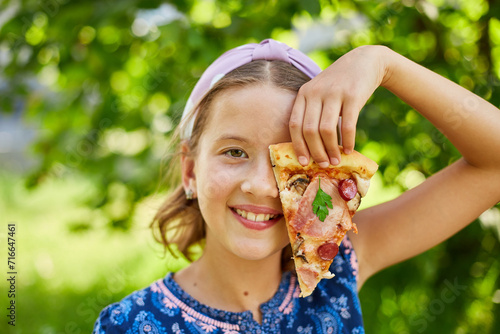 Cheerful Young Girl Enjoying a Slice of Pepperoni Pizza Outdoors on a Sunny Day