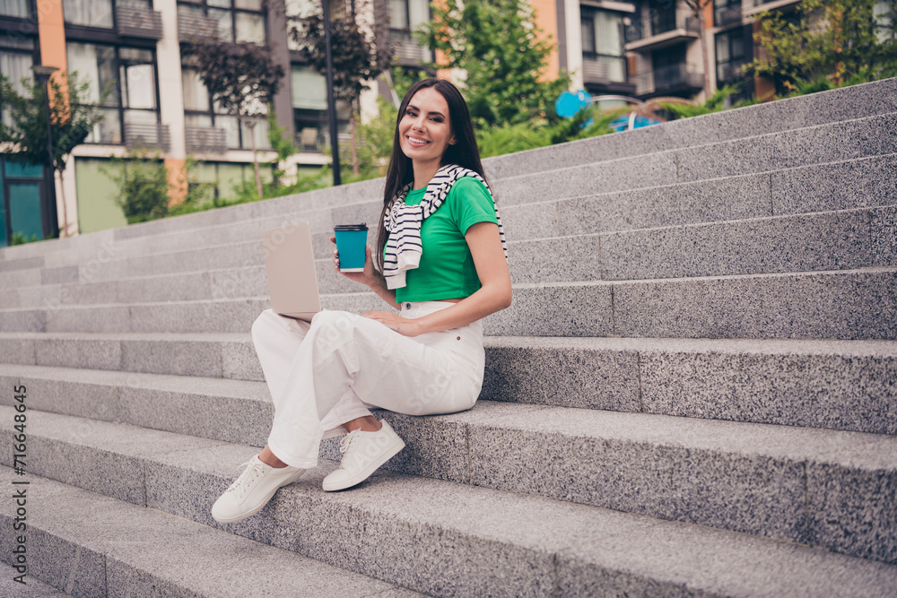 Fototapeta premium Photo of shiny sweet lady wear green t-shirt sitting stairs enjoying coffee communicating modern device outside urban city street