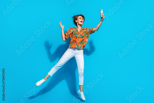 Full body photo of active excited lady jumping hold smart phone take selfie arm waving isolated on blue color background