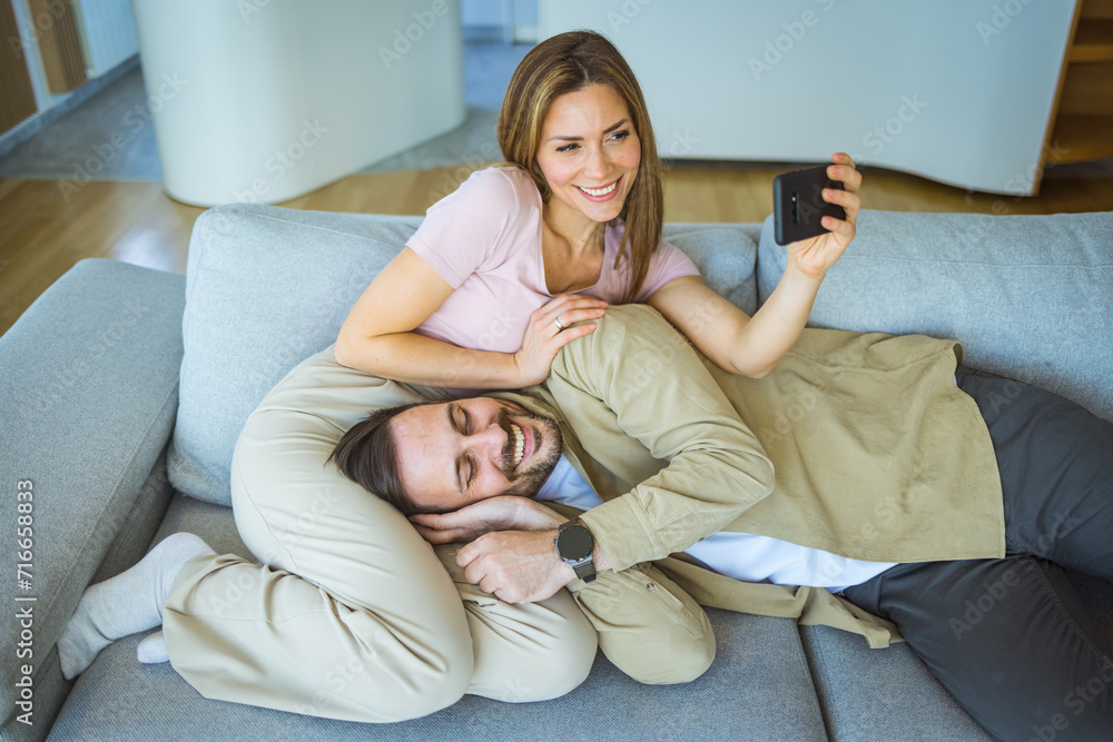 Man and woman couple make selfie by the smartphone sitting on sofa at home. Beautiful young loving couple bonding to each other and making selfie while sitting on the couch together Man and woman couple make selfie by the smartphone sitting on sofa at home. Beautiful young loving couple bonding to each other and making selfie while sitting on the couch together