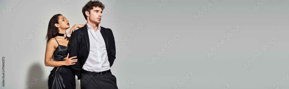 seductive young woman posing in black slip dress near handsome man in suit on grey backdrop, banner seductive young woman posing in black slip dress near handsome man in suit on grey backdrop, banner