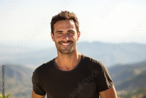 Portrait of a grinning man in his 30s dressed in a casual t-shirt against a panoramic mountain vista. AI Generation