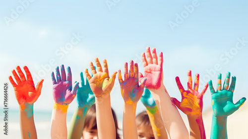children's hands in multi-colored paints stretch upward, against the backdrop of a blurred beach on a sunny day