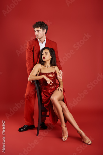 sophisticated young woman in sexy dress sitting on chair near man in formal wear on red background