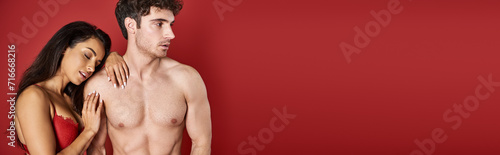 sensual young woman with brunette hair leaning on body of muscular man on red background, banner