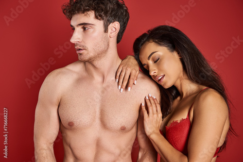 sensual young woman with brunette hair leaning on shirtless man on red background, affection