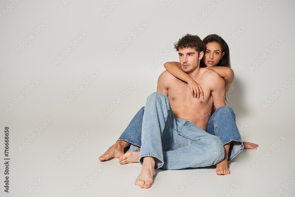 barefoot and sexy woman in blue jeans embracing her shirtless boyfriend on grey background, love