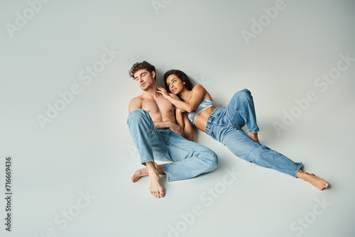 sexy young woman in silk bra leaning on handsome shirtless man in blue jeans on grey background