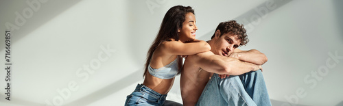 happy young woman in satin bra and jeans posing with shirtless man on grey background, banner