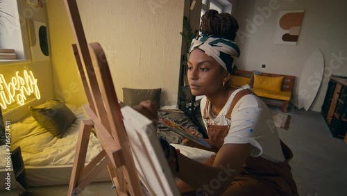 Side medium footage of female African American left-handed artist working on new painting using paintbrushes and palette in modern apartment photo