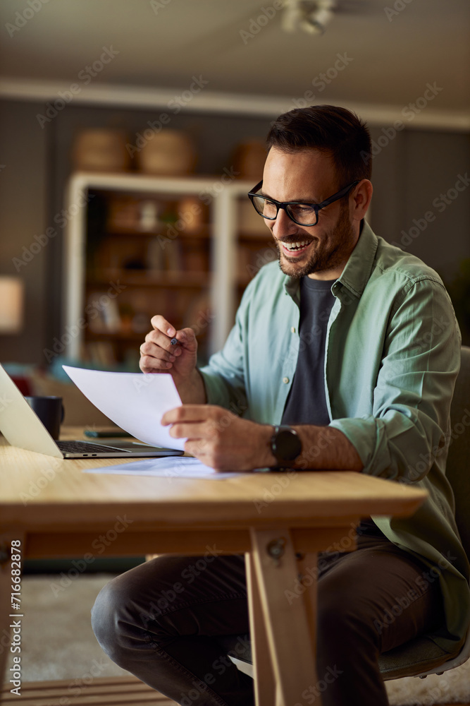 A happy adult male freelancer reading a document in his hand during an online meeting on a laptop.