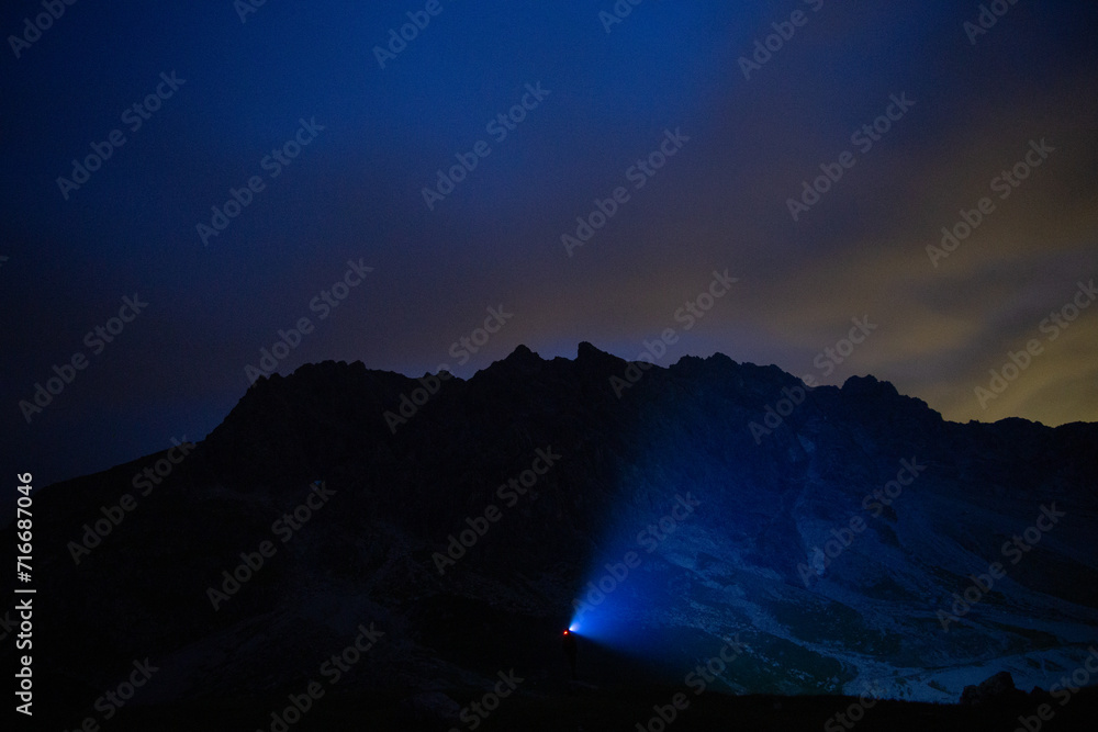 A lone hiker's headlamp illuminates the path in a mountainous terrain under a dusky sky.