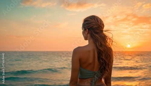  the back of a woman's head as she stands on the beach in front of the ocean with the sun setting behind her and the horizon in the distance.