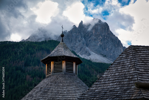 Blick auf die Spitze eines hölzernen Kirchturms mit Kreuz vor Bergpanorama mit in Wolken gehüllten Gipfeln