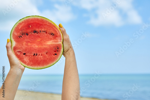 A persons hand is showcased against a serene beach backdrop, holding a ripe, juicy slice of watermelon.