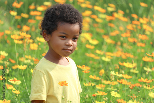 Portrait of happy smiling African boy with black curly hair standing around yellow orange cosmos flower garden. Kid spending time outdoors in beautiful meadow flower. Child has fun in summer park.