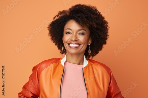 Portrait of a content afro-american woman in her 40s sporting a stylish varsity jacket against a pastel orange background. AI Generation