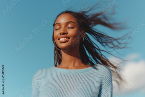 A young African American woman breathes deeply, enjoying a moment of the sun on her face outdoors on a sunny day with the blue sky in the background.
