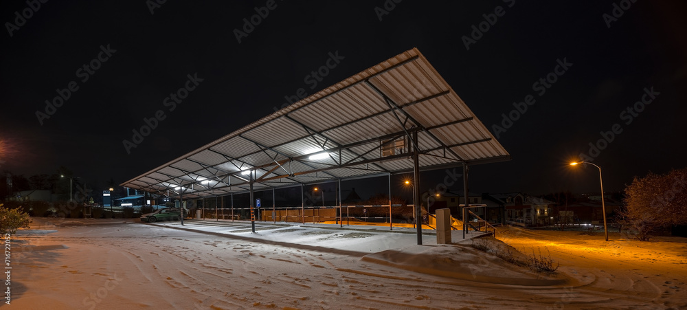 Wiata parkingowa oświetlona panelami słonecznymi - w śnieżną jesienną noc.Miasto (Ostrowiec) nocą - jasno oświetlony parking na tle białego śniegu. - obrazy, fototapety, plakaty 