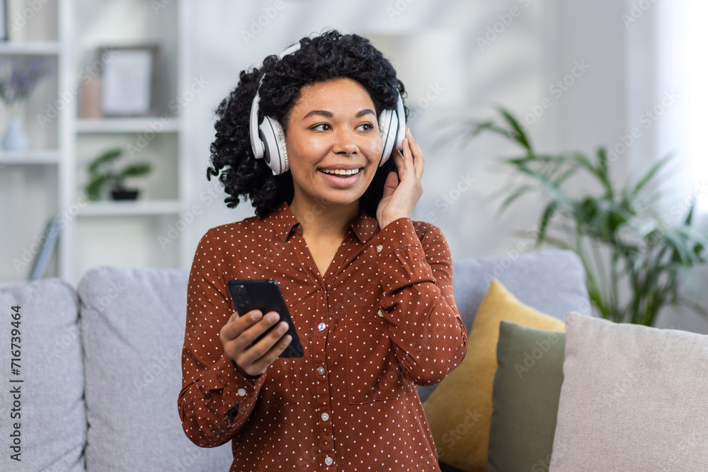 Close-up photo of a smiling and happy African American woman sitting on the couch at home wearing headphones and holding a mobile phone. Looks to the side Close-up photo of a smiling and happy African American woman sitting on the couch at home wearing headphones and holding a mobile phone. Looks to the side