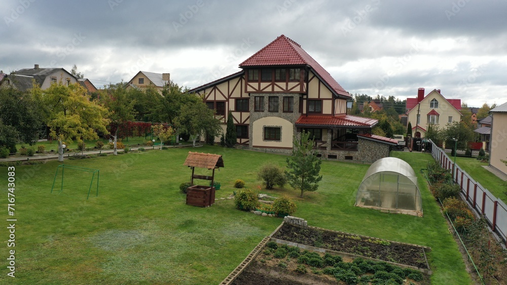Naklejka premium Large house, cottage with red tiled roof in English style with a plot against a gloomy sky. Green grass in front of the house in Europe. Soccer goal on a private plot.