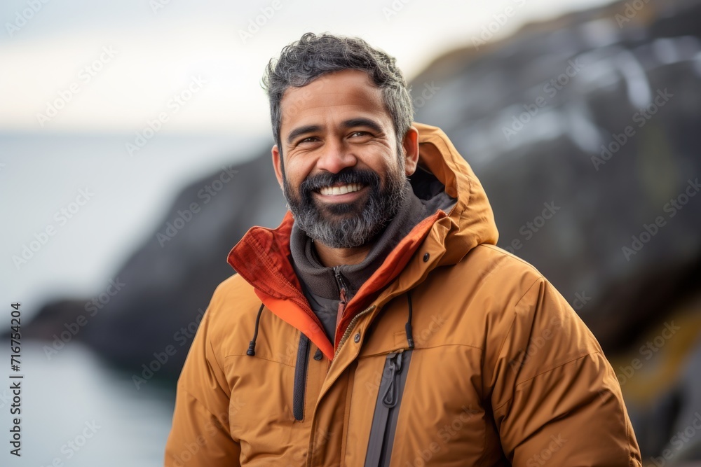 Portrait of a grinning indian man in his 40s donning a durable down jacket against a rocky shoreline background. AI Generation Portrait of a grinning indian man in his 40s donning a durable down jacket against a rocky shoreline background. AI Generation