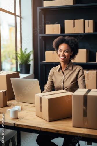 Warehouse serves as the backdrop to the seller's e-commerce empire. Smiley middle aged black woman sits in front of laptop monitor in a warehouse of products during online video call with a customer.