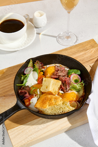 Sunny-side-up eggs in a skillet with roast beef, vegetables, and ciabatta, Americano coffee, and a glass of sparkling wine