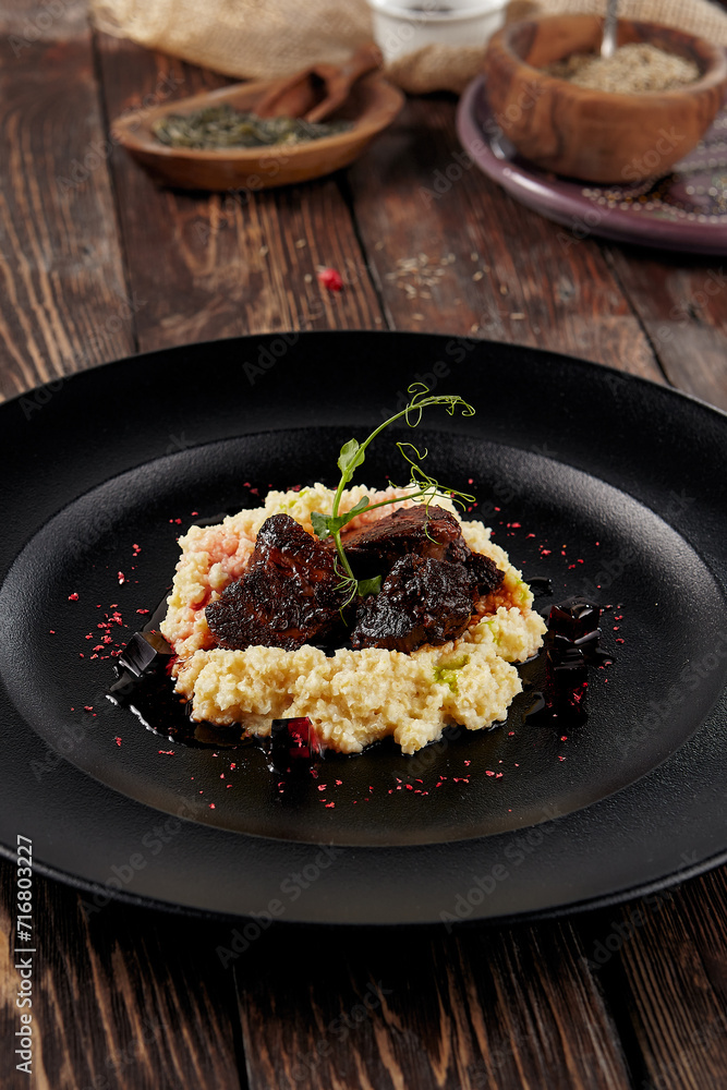 Braised beef cheeks with millet on a black plate, served on a rustic wooden table