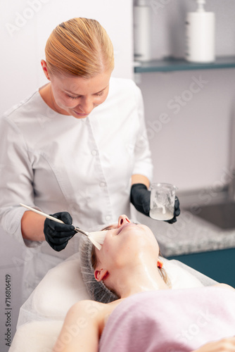 beautician in gloves applies a mask on the face of a client with a brush care for the skin