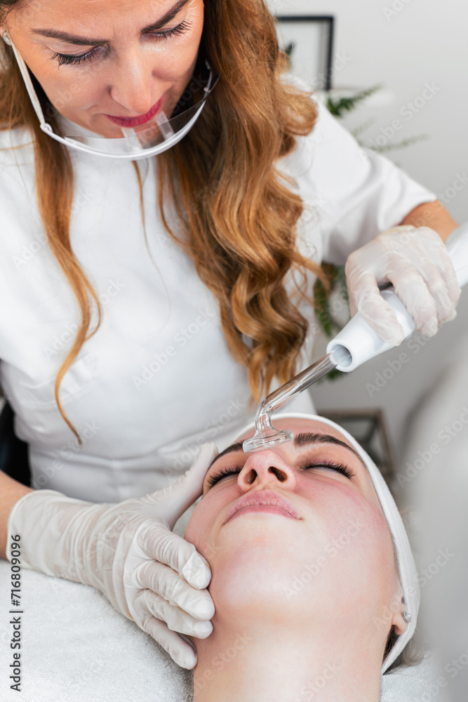 Young woman receiving face lift treatment with high-frequency wand. This modern treatment uses electric currents to kill acne-causing bacteria, reduce inflammation, and improve skin tone and texture. Young woman receiving face lift treatment with high-frequency wand. This modern treatment uses electric currents to kill acne-causing bacteria, reduce inflammation, and improve skin tone and texture.