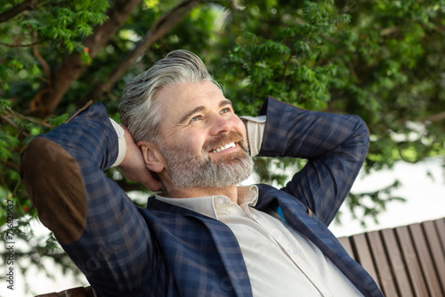 A 45 year old caucasian male in a dark blue blazer and white t-shirt, relaxing outdoors with a contented smile, exuding a serene and happy mood. © Liubomir