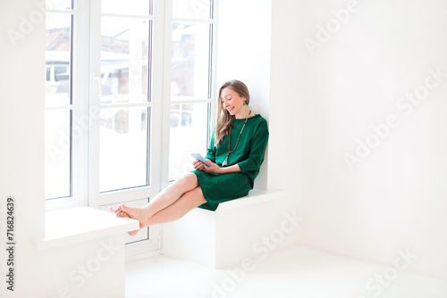 Young cheerful happy woman in dress holding mobile phone and smiling while sitting on windowsill