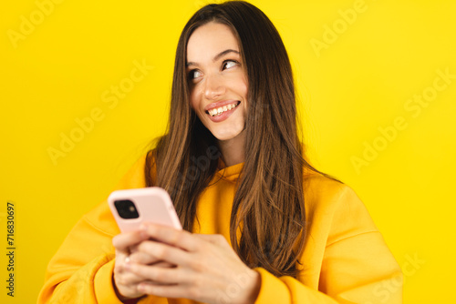 Smiling beautiful young woman in yellow hoody isolated on yellow background. Girl flirting while chatting on mobile phone, dreaming, look at side. Woman think about answer, look at side.