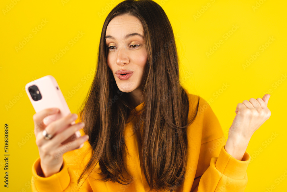 Fun young woman hold in hand use mobile cell phone doing winner gesture clenching fists isolated on yellow background studio. Girl use smartphone apps winning online celebrating discount gift voucher. Fun young woman hold in hand use mobile cell phone doing winner gesture clenching fists isolated on yellow background studio. Girl use smartphone apps winning online celebrating discount gift voucher.
