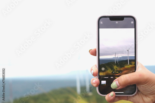 Woman hand using smartphone taking photo of windmill landscape view. Clean energy concept, wind power