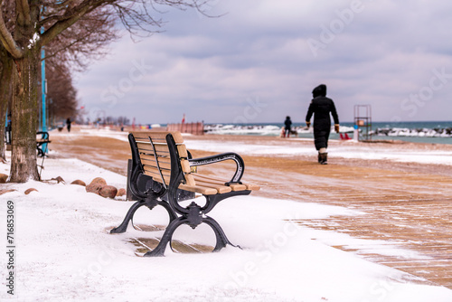 park bench on snowy winter day Kew Beach wooden boardwalk in Toronto's Beaches neigbourhood with dog walkers  (out of focus) in background room for text photo