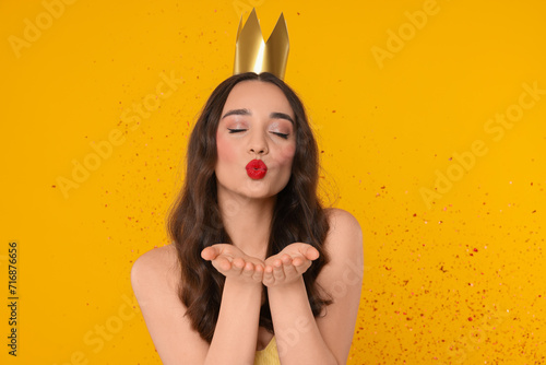 Beautiful young woman with princess crown blowing kiss under falling confetti on yellow background