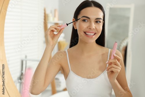 Beautiful young woman applying mascara near mirror in bathroom