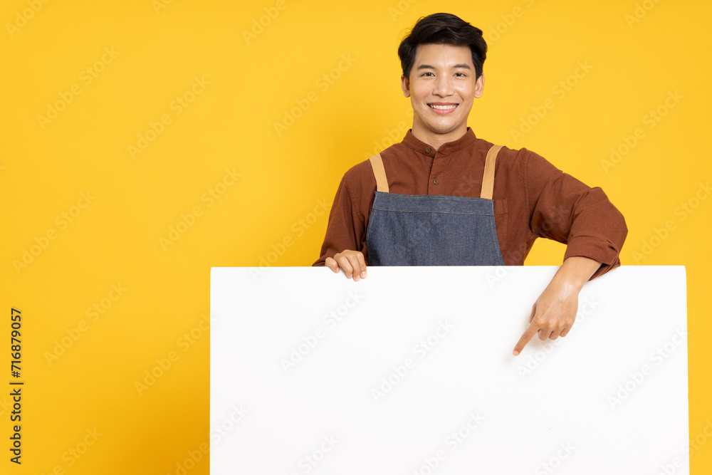 Restaurant owner sme young Asian man is pointing to white blank banner or empty copy space advertisement board isolated on yellow background, Looking at camera Restaurant owner sme young Asian man is pointing to white blank banner or empty copy space advertisement board isolated on yellow background, Looking at camera